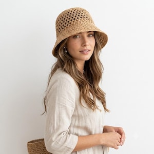 May include: A tan crochet bucket hat featuring an open diamond-shaped weave pattern on the crown and a solid, tightly woven brim. The hat is constructed from a natural-colored straw-like material with a rounded top and a downward-sloping brim.