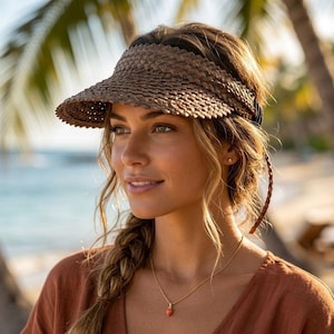 May include: A woven brown sun visor with a wide brim. The visor is made of a natural fiber material and has a dark band and braided ties. The person is wearing a gold necklace with a red pendant.