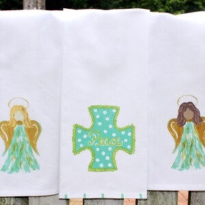 Puede incluir: Tres toallas de cocina blancas con dise&ntilde;os bordados. La primera toalla presenta un &aacute;ngel turquesa con alas doradas y un halo. La segunda toalla presenta una cruz turquesa con lunares blancos y la palabra "Paz" en escritura dorada. La tercera toalla presenta un &aacute;ngel de cabello casta&ntilde;o con alas doradas y un halo.
