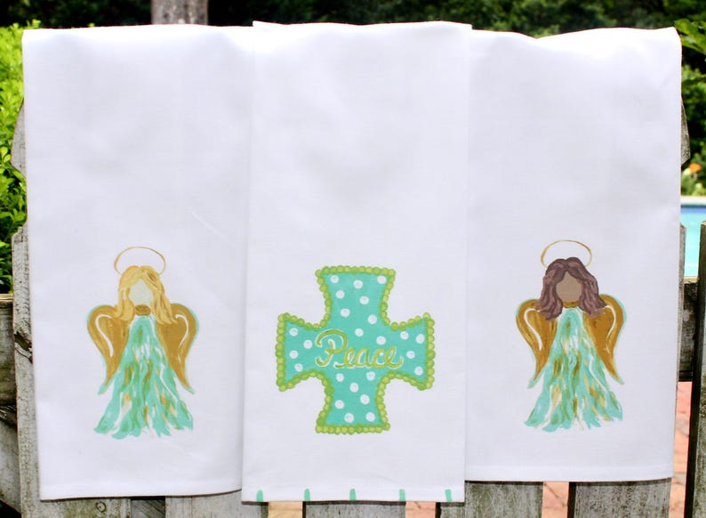 Puede incluir: Tres toallas de cocina blancas con dise&ntilde;os bordados. La primera toalla presenta un &aacute;ngel turquesa con alas doradas y un halo. La segunda toalla presenta una cruz turquesa con lunares blancos y la palabra "Paz" en escritura dorada. La tercera toalla presenta un &aacute;ngel de cabello casta&ntilde;o con alas doradas y un halo.
