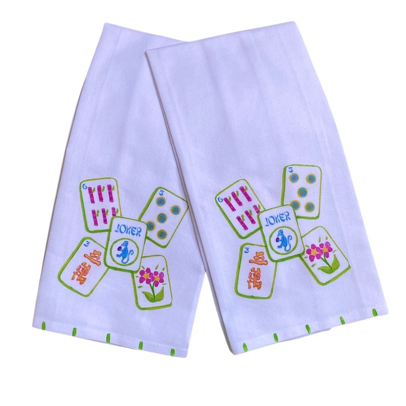Peut inclure: Deux torchons de cuisine blancs avec une bordure verte. Chaque torchon pr&eacute;sente un motif color&eacute; de dominos chinois avec un singe et le mot "JOKER" imprim&eacute; dessus.