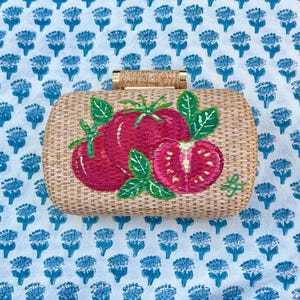 Puede incluir: Clutch rectangular de paja tejida con cierre dorado. La parte delantera presenta un dise&ntilde;o pintado a mano de tres tomates rojos con hojas verdes. El clutch descansa sobre una tela blanca con un estampado floral azul.