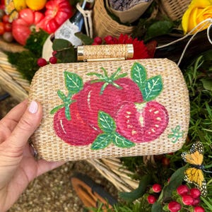 Puede incluir: Bolso de mano rectangular con textura de paja tejida. El bolso presenta un dise&ntilde;o pintado a mano de tomates rojos maduros con hojas verdes. El asa es un cilindro dorado. El fondo incluye vegetaci&oacute;n y una mariposa amarilla.