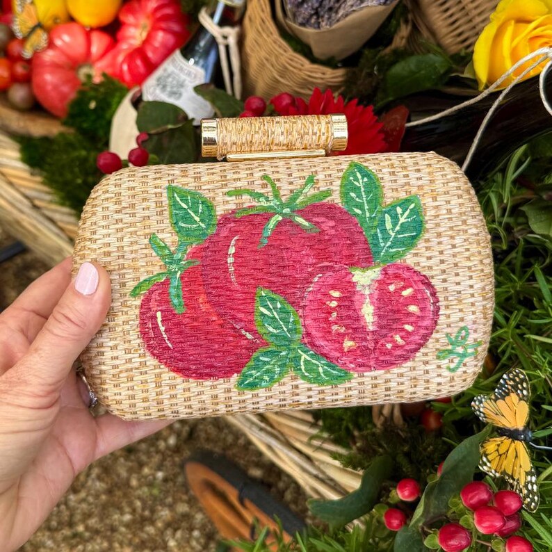 Puede incluir: Bolso de mano rectangular con textura de paja tejida. El bolso presenta un dise&ntilde;o pintado a mano de tomates rojos maduros con hojas verdes. El asa es un cilindro dorado. El fondo incluye vegetaci&oacute;n y una mariposa amarilla.