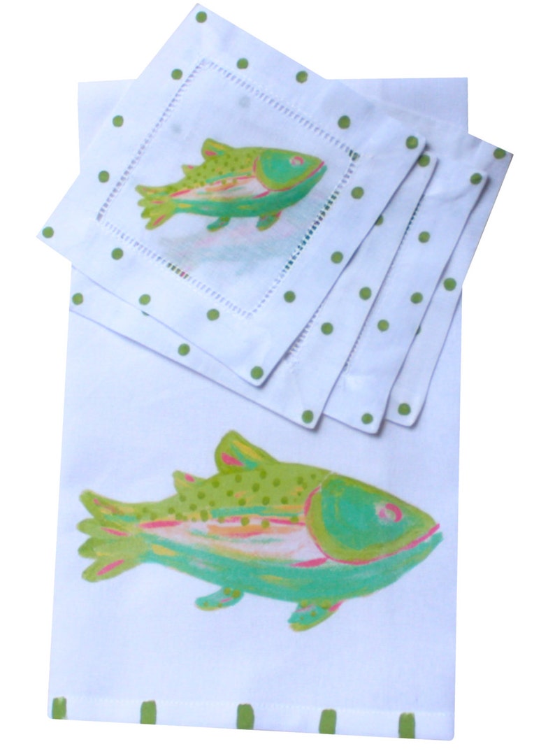 May include: Set of four white linen napkins with green polka dots and a green and pink fish design. The napkins are folded and stacked on top of a larger white linen napkin with a larger green and pink fish design.