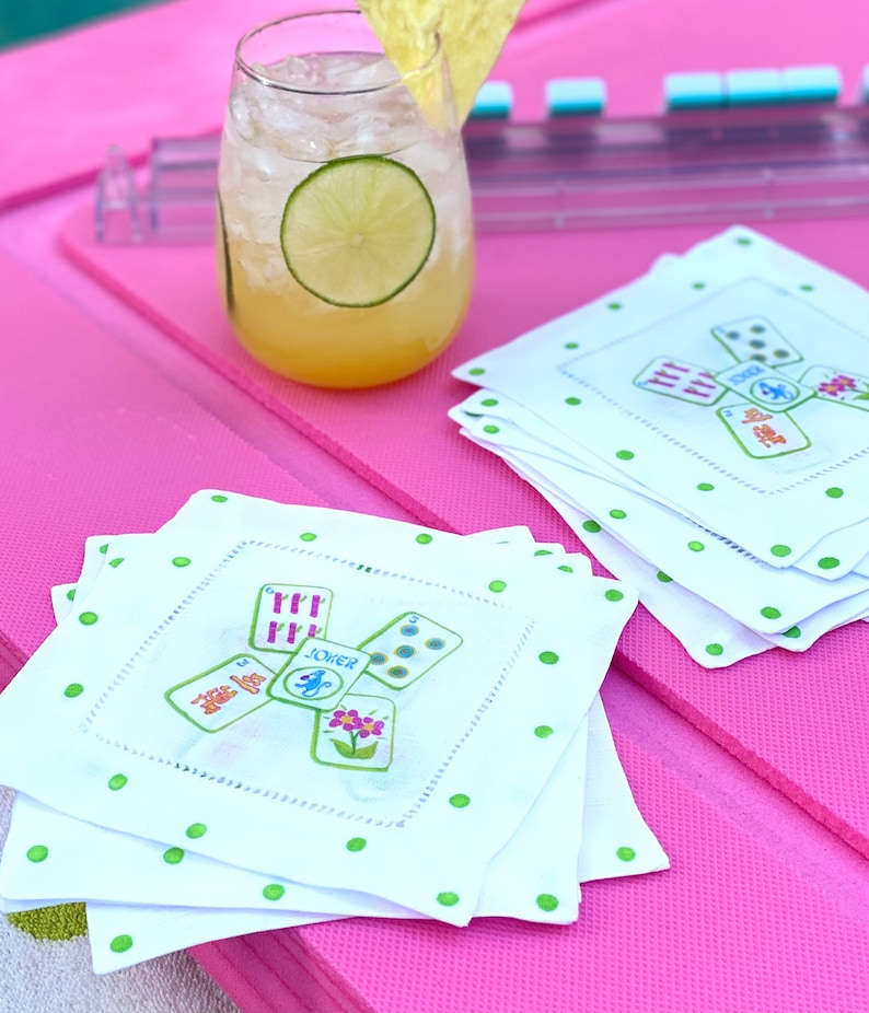 K&ouml;nnte beinhalten: Ein Stapel wei&szlig;er Leinen-Servietten mit gr&uuml;nen Punkten und einem bunten W&uuml;rfel-Design. Die Servietten zeigen das Wort "JOKER" und eine Blumengrafik. Die Servietten liegen auf einer rosa Oberfl&auml;che neben einem Glas mit einem gelben Getr&auml;nk und einer Limettenscheibe.