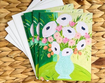 Könnte beinhalten: Ein Stapel weißer Karten und mehrere Kunstkarten mit Blumendesigns. Die Kunstkarten zeigen einen Strauß weißer Blumen mit schwarzen Zentren, rosa Blüten und orangefarbenen Akzenten in einer hellblauen Vase vor grünem Hintergrund.