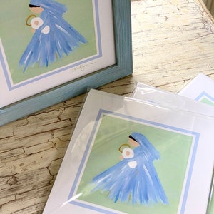 May include: A framed print of a painting depicting a woman in a blue dress holding a baby. The woman is wearing a blue veil and the background is a light green color. The print is signed "Shelly".