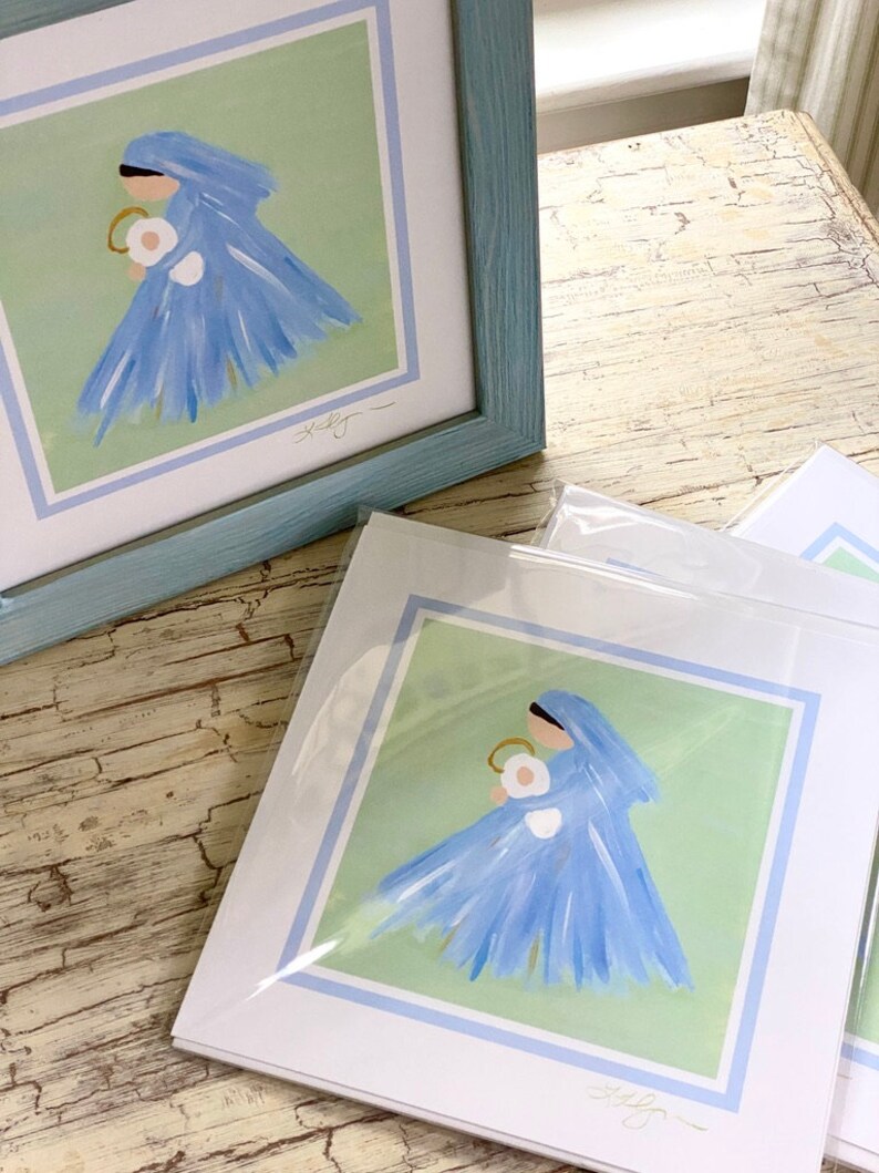 May include: A framed print of a painting depicting a woman in a blue dress holding a baby. The woman is wearing a blue veil and the background is a light green color. The print is signed "Shelly".