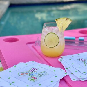 Peut inclure: Un cocktail rafra&icirc;chissant avec un morceau d'ananas et une tranche de citron vert dans un verre transparent. La boisson est sur une table rose avec des serviettes blanches &agrave; pois verts et un motif de cartes &agrave; jouer vert et blanc.