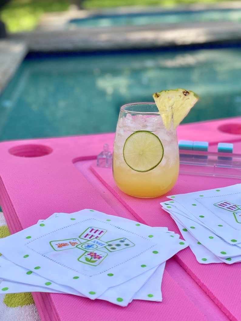Peut inclure: Un cocktail rafra&icirc;chissant avec un morceau d'ananas et une tranche de citron vert dans un verre transparent. La boisson est sur une table rose avec des serviettes blanches &agrave; pois verts et un motif de cartes &agrave; jouer vert et blanc.