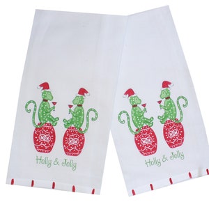 May include: Two white kitchen towels with a festive design. Each towel features a green monkey wearing a Santa hat and holding a cocktail, perched atop a red ornament. The words "Holly & Jolly" are printed below the monkeys.