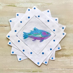May include: Set of four white linen napkins with blue polka dots and a colorful fish design. The napkins are folded and stacked on a white wooden surface.