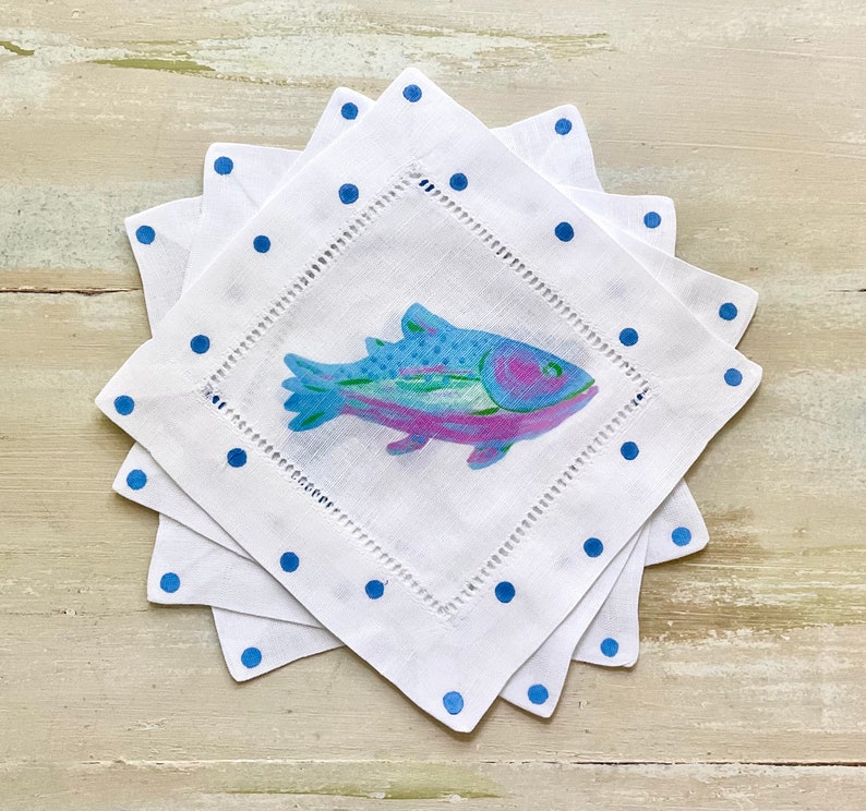 May include: Set of four white linen napkins with blue polka dots and a colorful fish design. The napkins are folded and stacked on a white wooden surface.