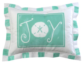 Pode incluir: Almofada retangular branca com um centro turquesa com a palavra "JOY" em uma fonte extravagante e um desenho de dólar de areia branco. A almofada tem uma borda branca com listras turquesas e uma costura decorativa.