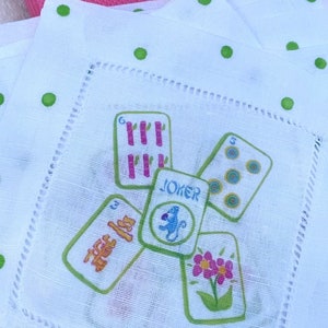 K&ouml;nnte beinhalten: Wei&szlig;e Cocktail-Serviette aus Leinen mit gr&uuml;nen Punkten und einem Design aus Spielkarten mit einem Joker in der Mitte. Die Karten sind mit Zahlen und Symbolen bedruckt.