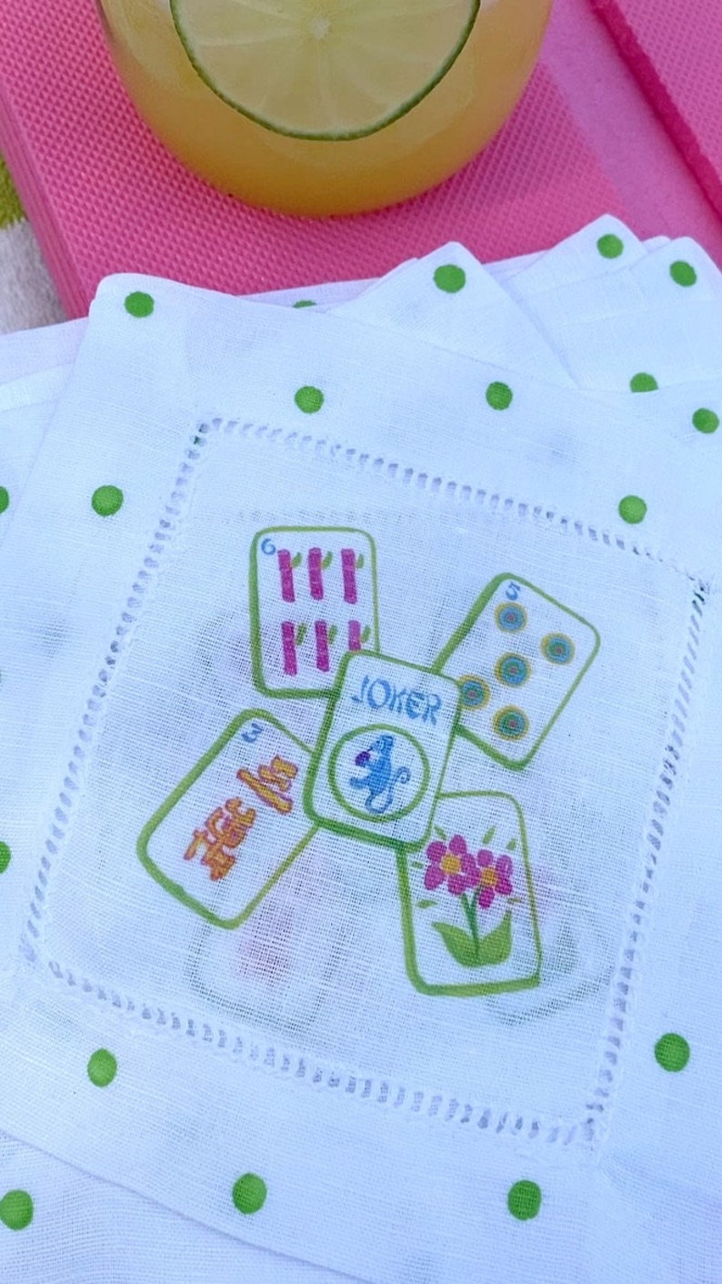 K&ouml;nnte beinhalten: Wei&szlig;e Cocktail-Serviette aus Leinen mit gr&uuml;nen Punkten und einem Design aus Spielkarten mit einem Joker in der Mitte. Die Karten sind mit Zahlen und Symbolen bedruckt.