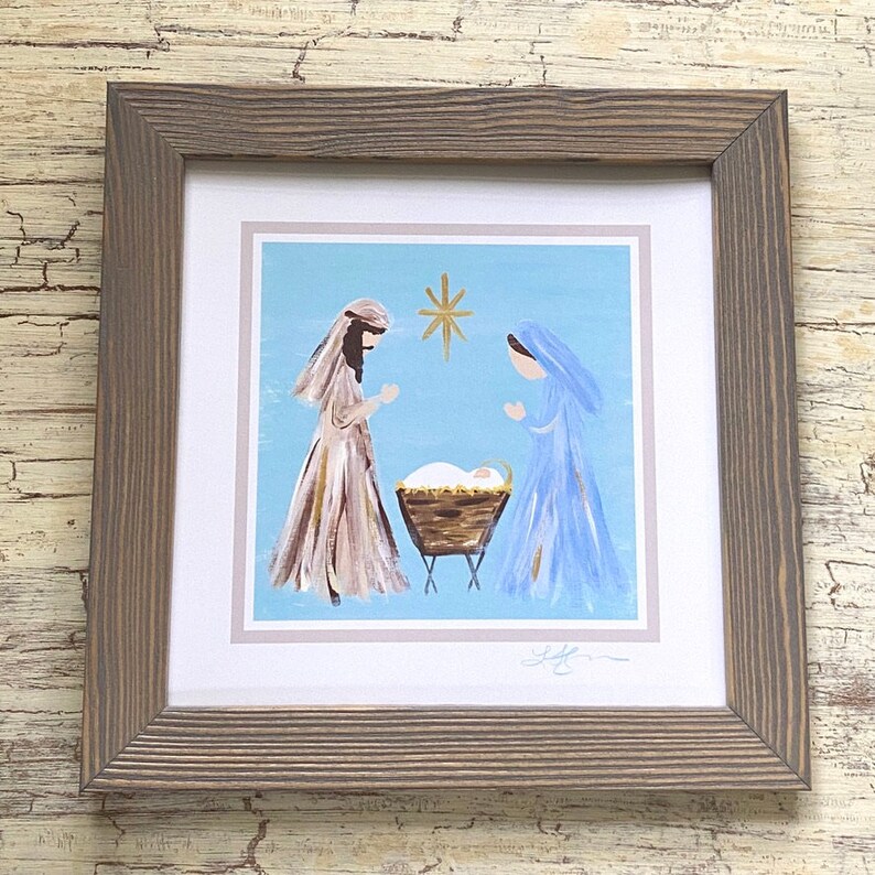 May include: A framed painting depicting the nativity scene with Mary and Joseph standing beside a manger with a baby inside. The painting is done in a simple, folk art style with a blue background and a gold star above the manger.