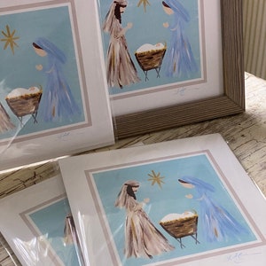 May include: A set of four framed prints depicting a nativity scene with Mary, Joseph, and baby Jesus in a manger. The prints are on a light blue background with a gold star above the manger. The prints are signed by the artist.