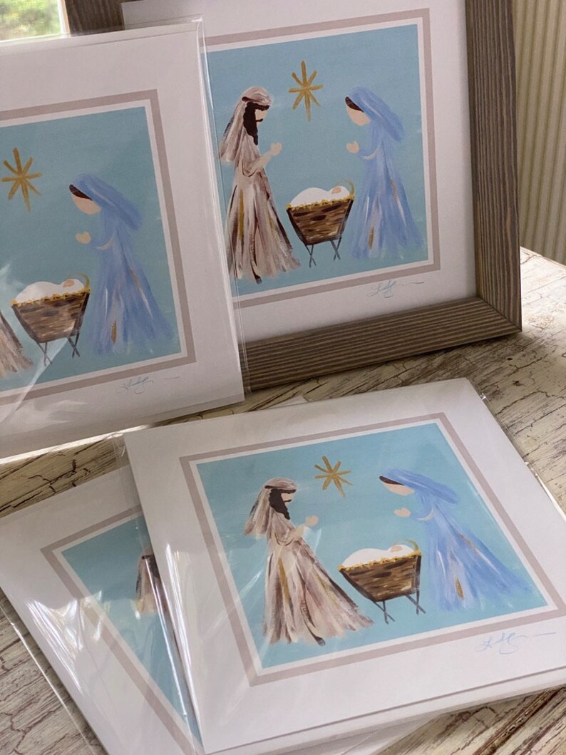 May include: A set of four framed prints depicting a nativity scene with Mary, Joseph, and baby Jesus in a manger. The prints are on a light blue background with a gold star above the manger. The prints are signed by the artist.