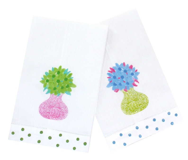 May include: Two white kitchen towels with green and blue polka dot borders. One towel features a pink vase with green flowers and the other features a green vase with blue flowers.