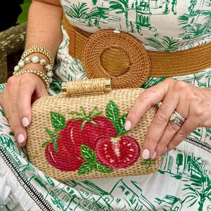Puede incluir: Bolso de mano rectangular tejido con un dise&ntilde;o de tomates rojos y hojas verdes. El bolso tiene un cierre dorado. El fondo incluye un cintur&oacute;n tejido y un vestido con estampado verde y blanco. La persona lleva pulseras y anillos dorados.