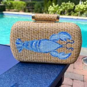 May include: A rectangular woven straw clutch bag featuring a painted light blue lobster graphic on the front. The bag has a cylindrical woven straw top handle with gold-tone metal accents at each end, providing a structured and compact design.