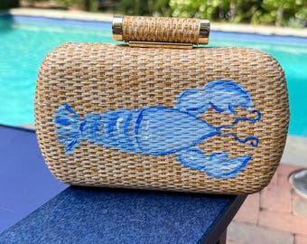 Puede incluir: Un bolso de mano rectangular de paja tejida con un gráfico de langosta azul claro pintado en la parte delantera. El bolso cuenta con un asa superior cilíndrica de paja tejida con detalles metálicos en tono dorado en cada extremo.