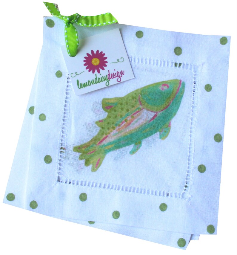 May include: Set of four white linen napkins with green polka dots and a green and blue fish design. The napkins are trimmed with white stitching. A green ribbon with a white polka dot pattern is tied around the napkins with a tag that reads "lemondaisydesign".