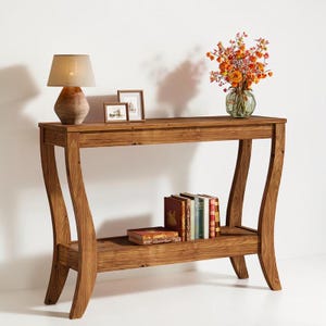 May include: A wooden console table with a lower shelf, holding books and a decorative lamp. The table has curved legs and a natural wood finish. A vase of orange flowers and framed pictures are on top.