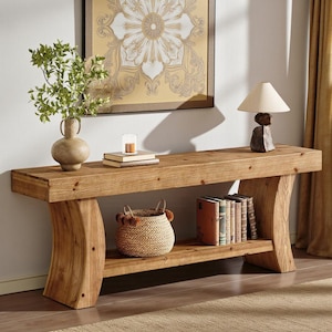 May include: A long wooden console table with a natural grain finish and curved legs. The top surface holds a beige ceramic vase with green foliage, a small candle, and two stacked books. A woven basket and several books sit on the lower shelf.