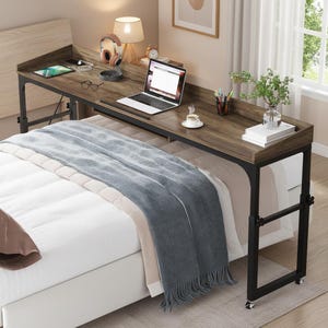 May include: A brown and black rolling desk with a laptop, headphones, and a tablet on top. The desk is positioned over a bed with a blue blanket. A lamp and a small clock are also visible.