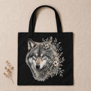 Op de afbeelding: Een zwarte draagtas met een gedetailleerde illustratie van een grijze wolvenkop op de voorzijde. De wolf is gedeeltelijk omringd door een cluster van witte bloemen en bladerrijke botanische elementen, wat een natuurlijk contrast vormt.