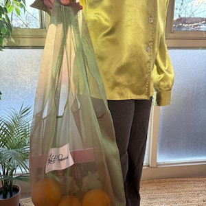 May include: An olive green mesh tote bag filled with oranges and books. The bag has a white label with the text "Hot Pieces". The oranges are bright orange. The person is wearing a light green satin shirt and dark grey trousers.