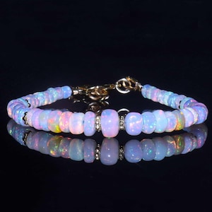 Può includere: Un braccialetto di opale con chiusura dorata. Il braccialetto presenta perle di opale ovali e iridescenti nei toni del blu, rosa e giallo. Le perle sono infilate, creando un effetto scintillante.