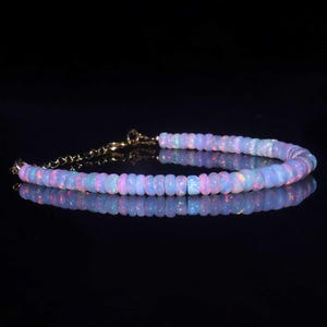 May include: A delicate bracelet featuring iridescent opal beads. The beads are a soft white color with flashes of pink, blue, and green. The bracelet is strung on a gold-colored chain and clasp. The jewelry is displayed on a reflective black surface.