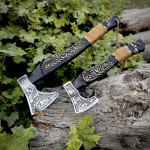May include: Two decorative axes with intricate carvings. The axe heads are silver-colored with detailed designs, while the handles are black with brown twine wrapping. The axes are displayed on a weathered wooden surface, surrounded by green foliage.