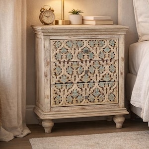 Puede incluir: Una mesita de noche blanca envejecida con tres cajones con un diseño floral tallado en azul claro. Una lámpara dorada, un despertador y una pequeña planta están encima. La mesita está junto a una cama con cabecero y ropa de cama de color neutro.