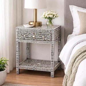 Może przedstawiać: Czarno-biały stolik nocny z kwiatowym wzorem, z szufladą i półką. Stolik nocny ma złote okucia i jest zwieńczony białą lampą i wazonem z białymi kwiatami. Stolik nocny ma około 61 cm wysokości.