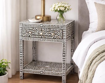 Può includere: Comodino con motivo floreale bianco e nero, con cassetto e ripiano. Il comodino ha ferramenta color oro ed è sormontato da una lampada bianca e un vaso di fiori bianchi. Il comodino è alto circa 61 cm.