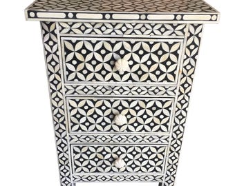 Può includere: Un comodino a fantasia bianca e nera con tre cassetti. Il comodino presenta un design geometrico sulla parte superiore, sui lati e sui frontali dei cassetti. Ogni cassetto ha una manopola bianca a forma di fiore. Il comodino è alto circa 61 cm.