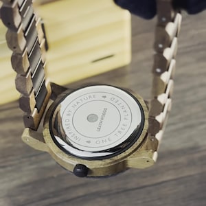 May include: Close-up of a wooden watch with a light brown band and face. The watch back is visible, displaying the text "INSPIRED BY NATURE" and "ONE TREE PLANTED". The watch has a circular face and a dark button.
