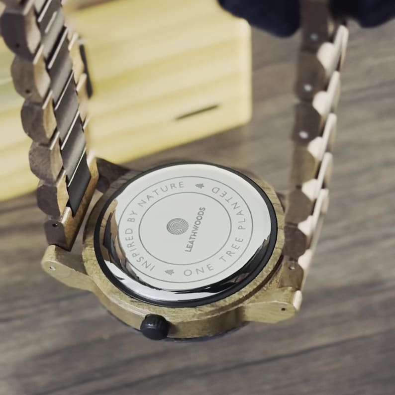 May include: Close-up of a wooden watch with a light brown band and face. The watch back is visible, displaying the text "INSPIRED BY NATURE" and "ONE TREE PLANTED". The watch has a circular face and a dark button.