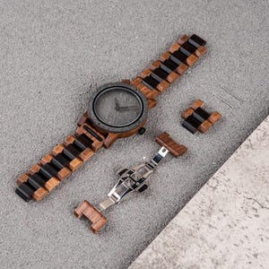 May include: A wooden wristwatch with a dark gray face and a band made of alternating dark and light wood segments. The watch is accompanied by a detached band segment, a clasp, and two small pins, all on a gray surface.