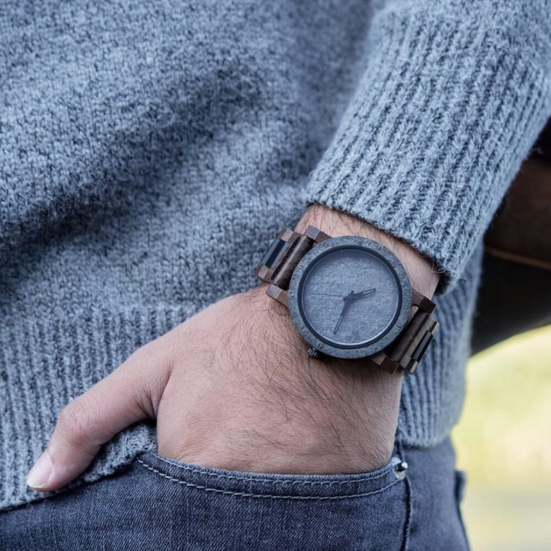May include: A wooden watch with a dark gray face and a matching wooden band is worn on a wrist. The watch has a minimalist design with Roman numeral hour markers. The watch is paired with a gray sweater and blue jeans.