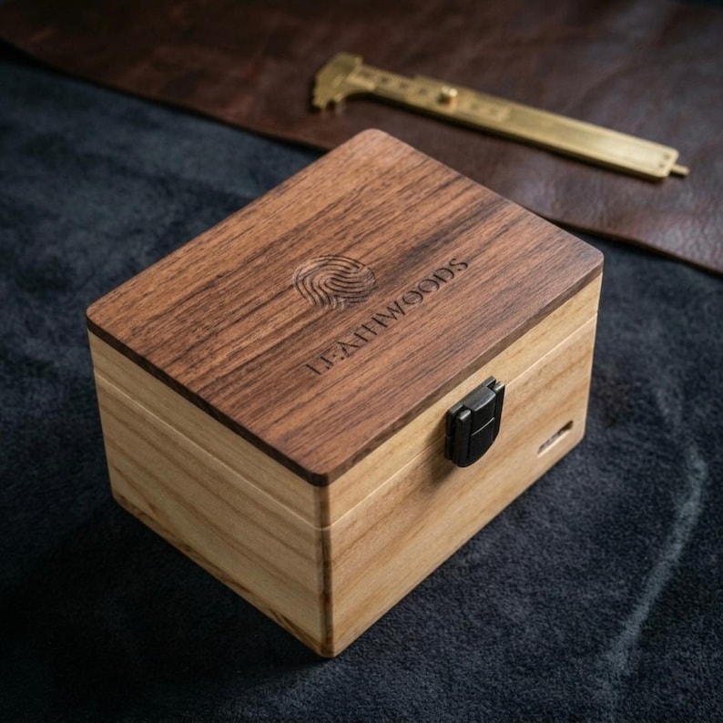 May include: A rectangular wooden box with a dark wood lid and a lighter wood body. The lid is engraved with the text "LEATHWOODS" and a fingerprint design. A black clasp secures the box. A brass caliper is in the background.