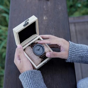 May include: A wooden watch with a dark gray face and brown band rests in a light-colored wooden box. The watch is displayed on a beige cushion inside the box. The box is open, revealing the watch and its presentation.