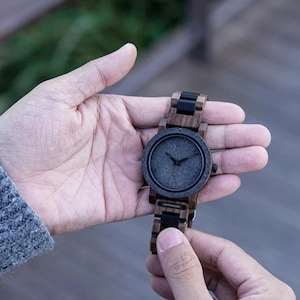 May include: A wooden wristwatch with a dark gray face and black hands. The watch has a wooden band with black accents. The watch is held in a person's hand.