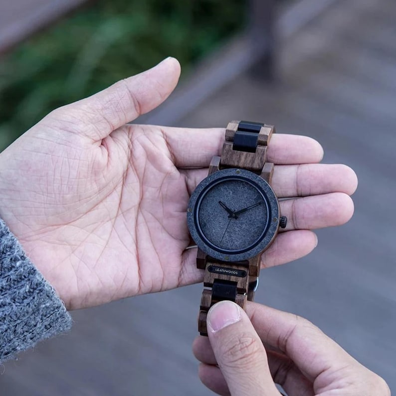 May include: A wooden wristwatch with a dark gray face and black hands. The watch has a wooden band with black accents. The watch is held in a person's hand.