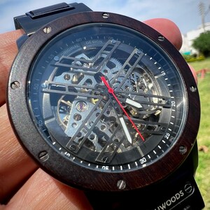 May include: A skeleton watch with a dark wood bezel and black band. The watch face reveals the intricate inner workings with silver gears and a red second hand. The watch is marked "ORIGINAL DESIGN" and "LEATHWOODS".